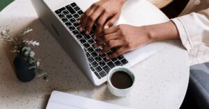 A person typing on a laptop at a café table with coffee and a notebook.